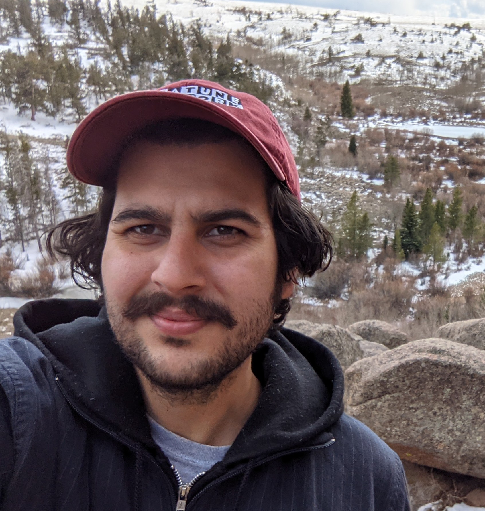 Selfie of a man wearing a hat standing against a wide open mountain background.