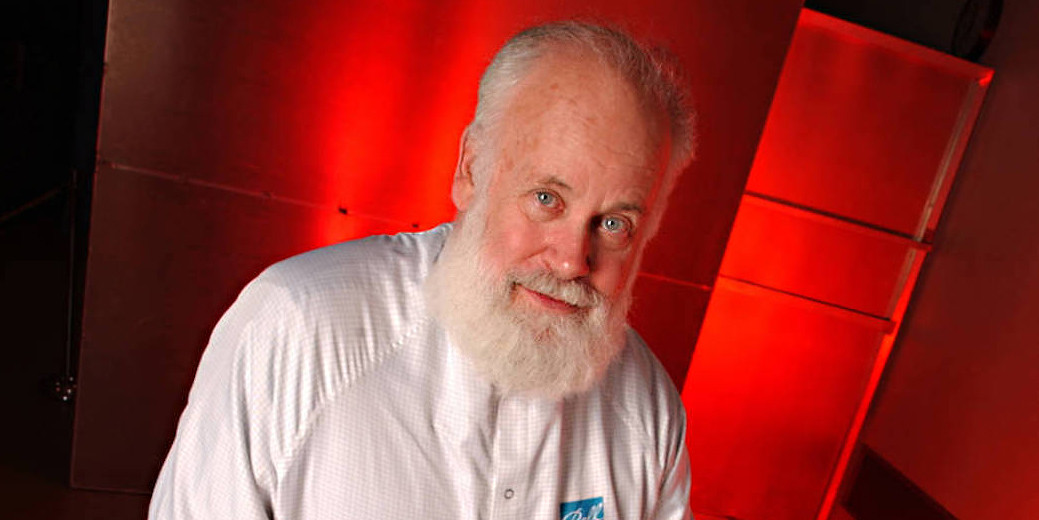 Professor Michael A'Hearn, an older man with a full white beard, in front of a red background. Photo by NASA.