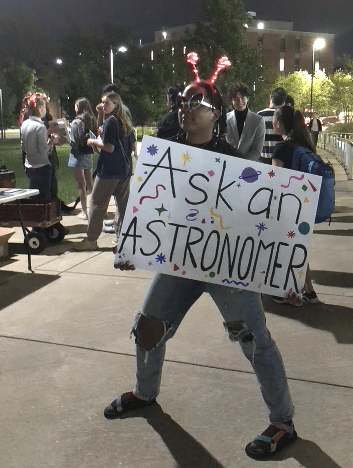 Grad student and event organizer Jordan wearing crazy sunglasses and light-up headband antennae and holding an Ask an Astronomer sign
