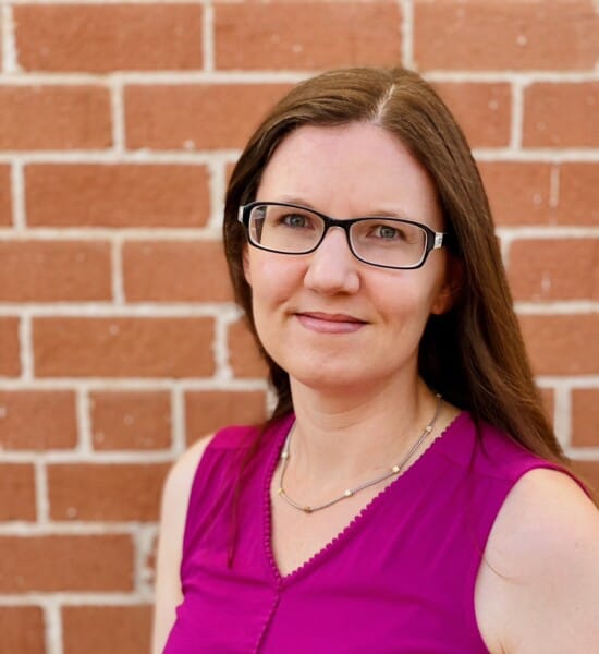 Kat Volk; woman with long brown hair standing in front of a brick wall