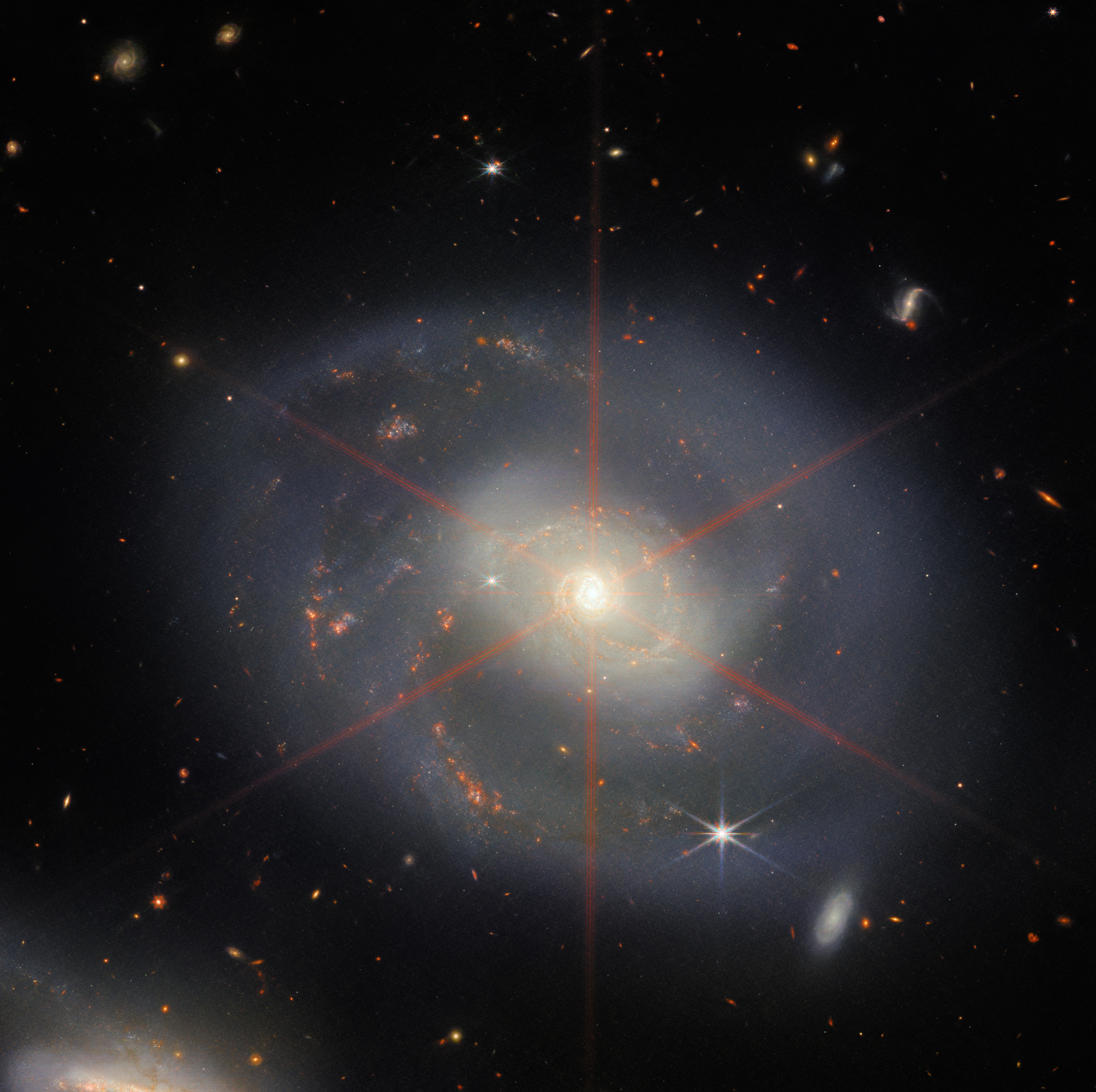 JWST image of spiral galaxy NGC 7469, featuring a white core that emits red six-pointed lines across its center. Faint-blue spiral arms dotted with orange-red dots sit against the dark background of space.
