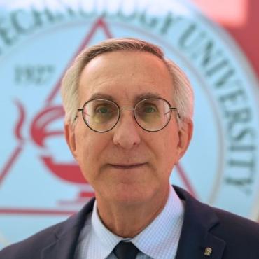 white-haired man wearing glasses in front of Capitol Tech Univ logo