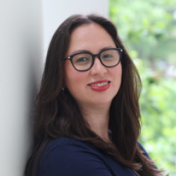 young woman with long dark brown hair and glasses leaning against white wall outside