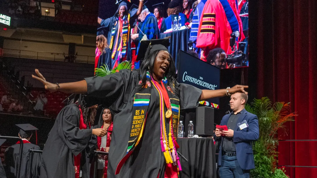 A female Bachelor's degree recipient in colorful regalia celebrates as she crosses the graduation stage.