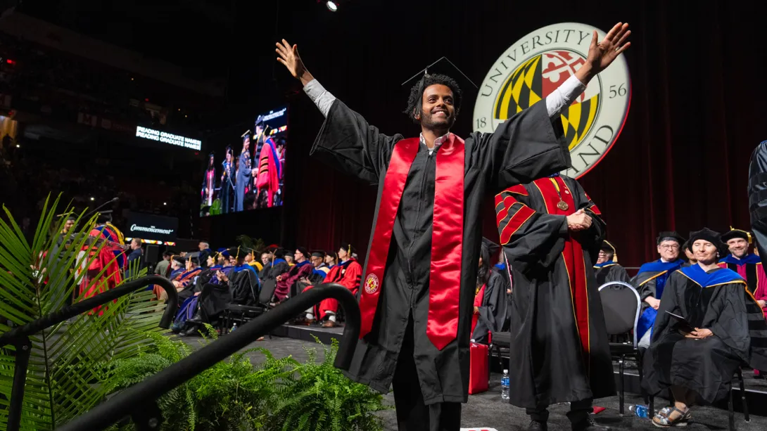 A male Bachelor's degree recipient celebrates by throwing up his arms as he crosses the graduation stage.